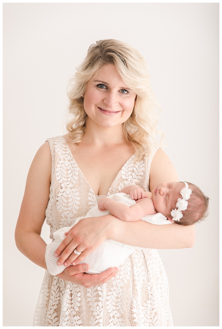 mother styled with curly blonde hair and makeup wearing lulus lace dress holds newborn baby girl by meghan goering photography
