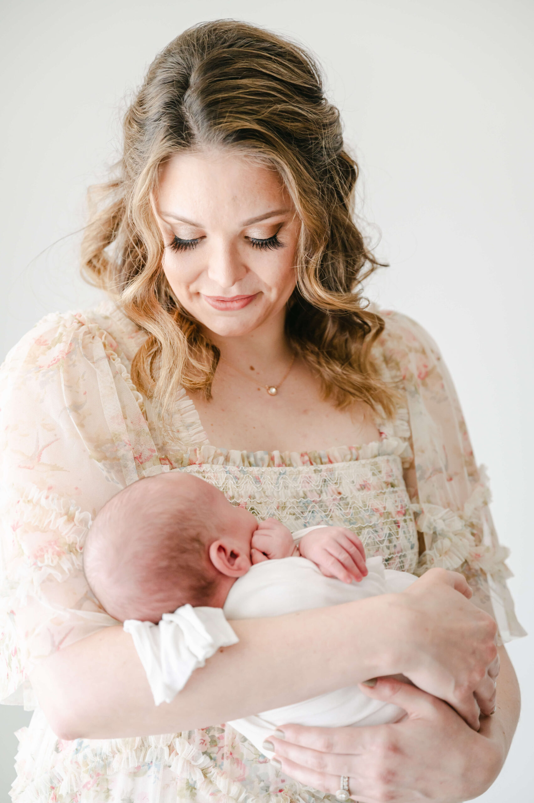 gorgeous first time mama wearing needle and thread smocked avery dress. She has professional hair and makeup with stunning eyes and fake lashes and is holding her newborn baby boy