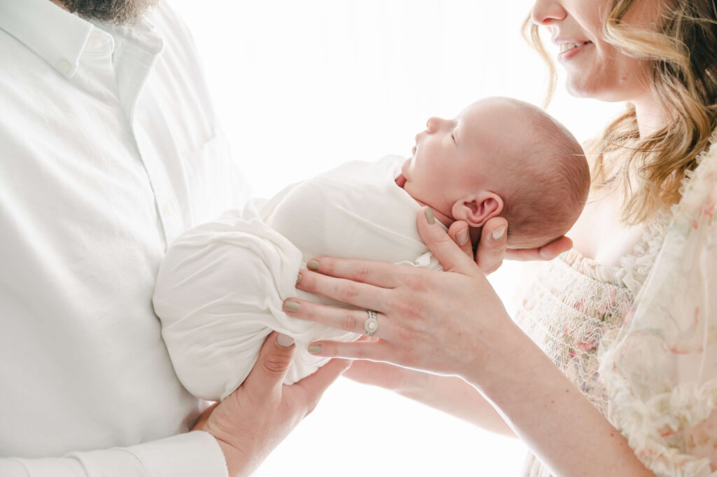 beautiful couple in light and airy photography studio holding newborn baby with backlighting